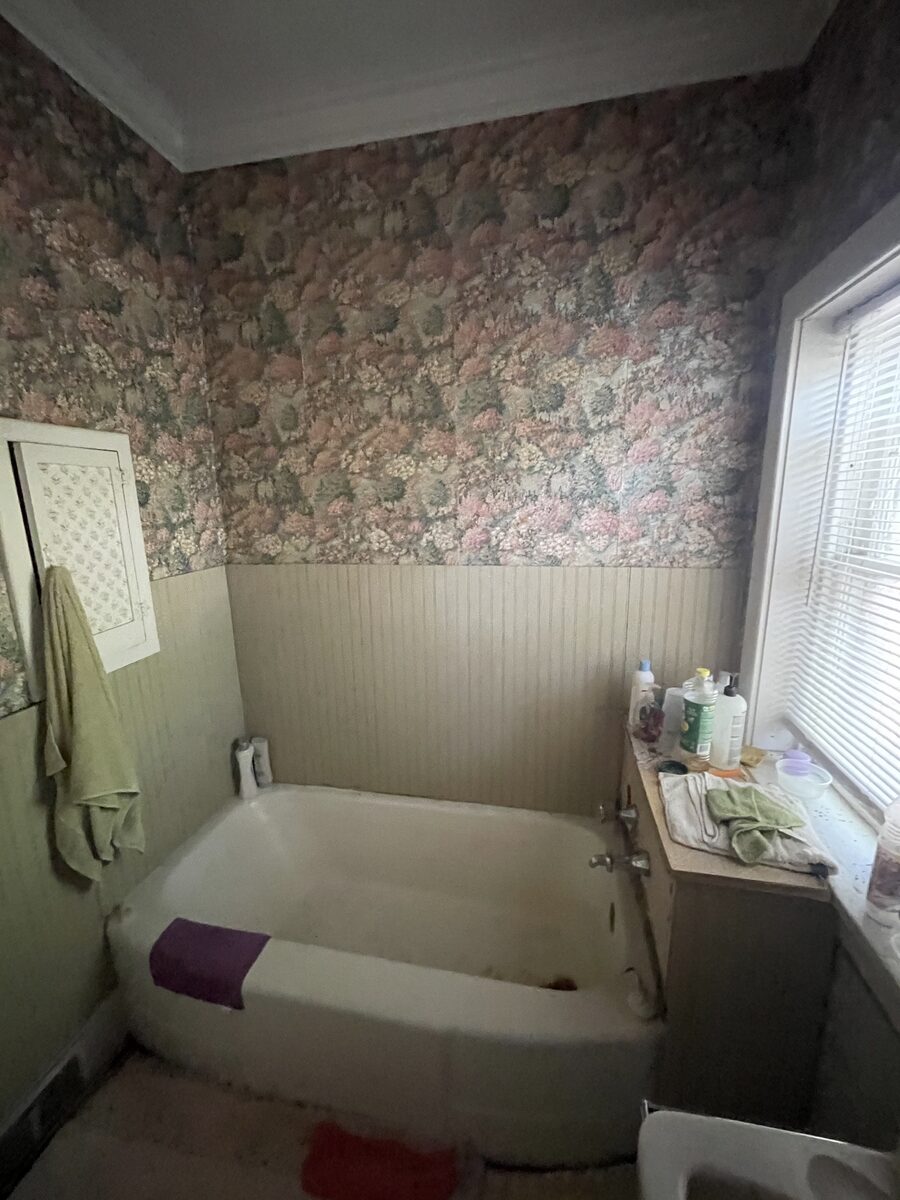 The home's original bathroom with dated floral wallpaper and worn fixtures